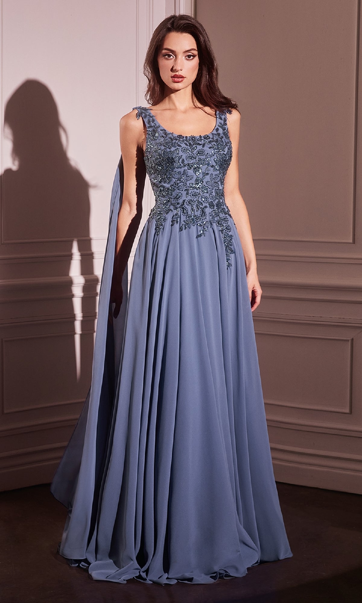 A woman stands indoors wearing the Ladivine CDS509 Shoulder Sash Long Formal Dress, a floor-length, sleeveless blue gown with floral embroidery on the bodice and a flowing skirt.
